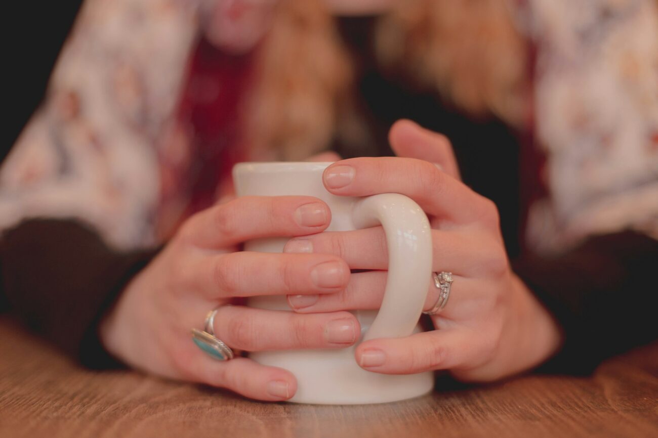 Two hands wrapped around a white mug on a wooden table to warm up