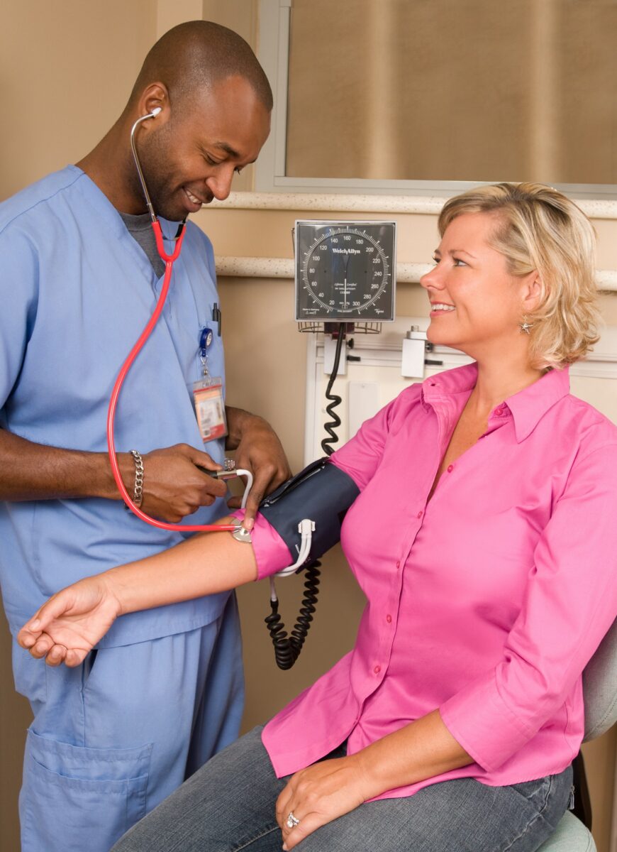 A nurse in blue scrubs checking a smiling woman's blood pressure in a clinic