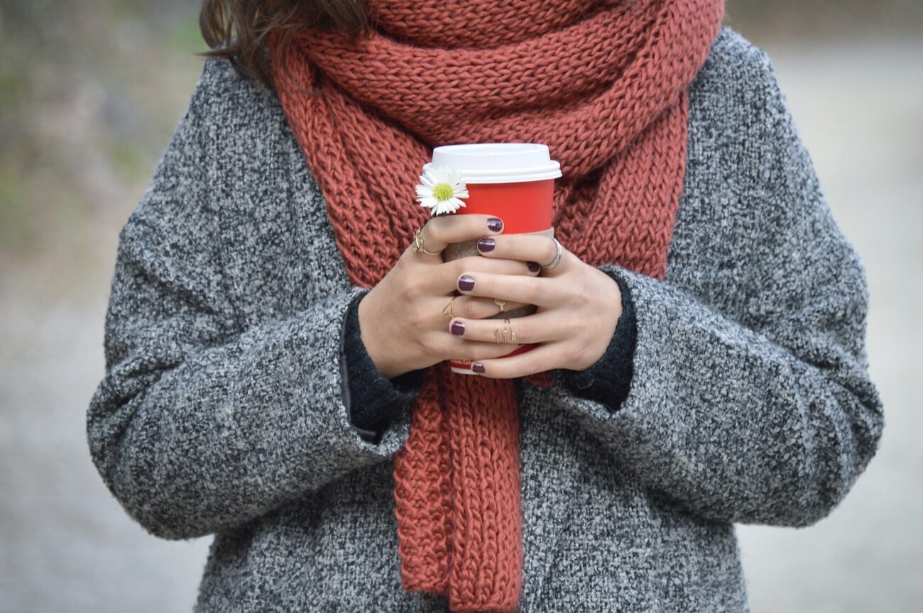 A woman in a wool coat and scarf holding a hot drink in both cold hands outdoors